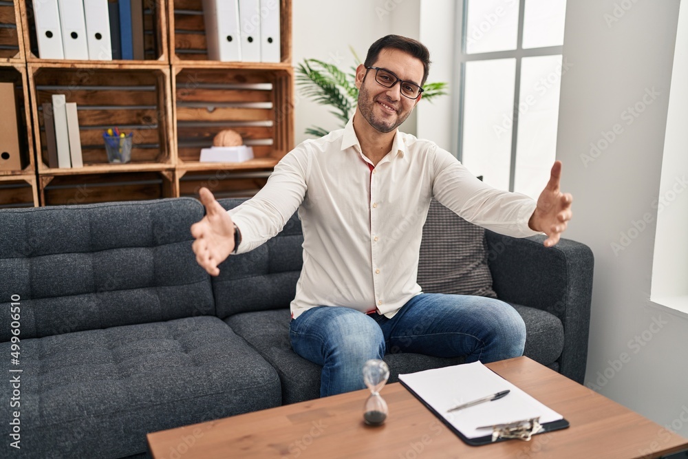 Young hispanic man with beard working at consultation office looking at the camera smiling with open arms for hug. cheerful expression embracing happiness. Young hispanic man with beard working at consultation office looking at the camera smiling with open arms for hug. cheerful expression embracing happiness.