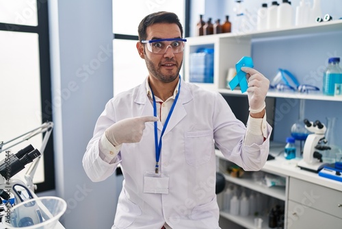 Young hispanic man with beard working at scientist laboratory holding blue ribbon pointing finger to one self smiling happy and proud