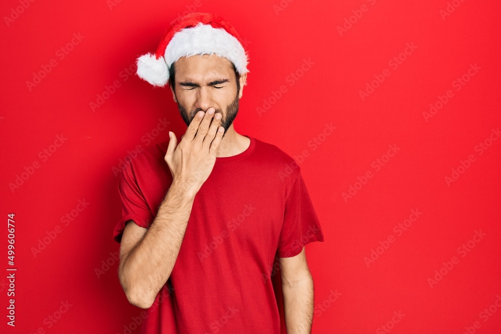 Young hispanic man wearing christmas hat bored yawning tired covering mouth with hand. restless and sleepiness.