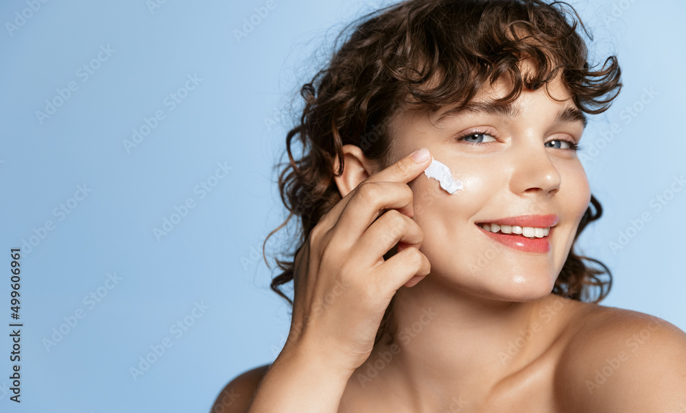Obraz premium Close up portrait of smiling young woman with curly hair, using facial skincare cream on her face, doing cosmetic daily routine after shower, standing over blue background