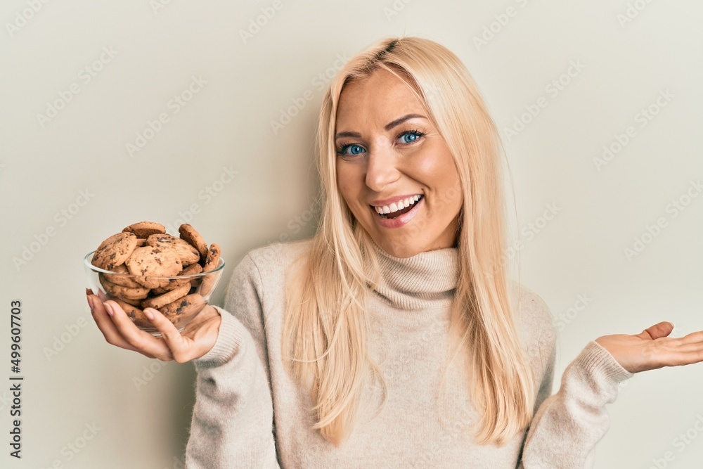 Young blonde woman holding bowl with chocolate chips cookies celebrating achievement with happy smile and winner expression with raised hand Young blonde woman holding bowl with chocolate chips cookies celebrating achievement with happy smile and winner expression with raised hand