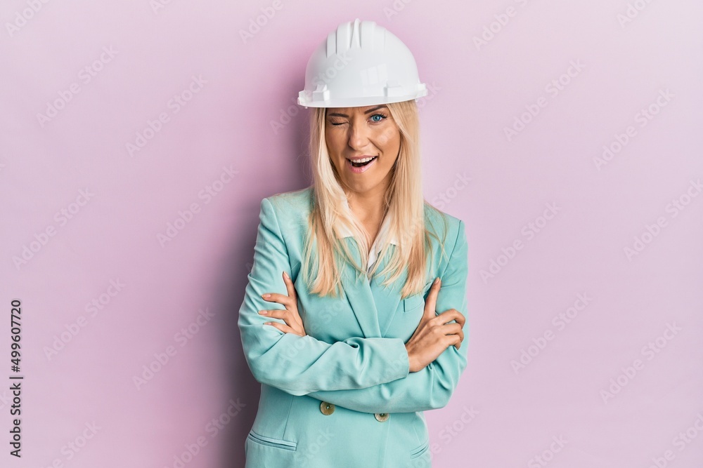 Young blonde woman wearing architect hardhat winking looking at the camera with sexy expression, cheerful and happy face.