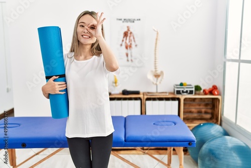 Asian young woman holding yoga mat at pain recovery clinic smiling happy doing ok sign with hand on eye looking through fingers