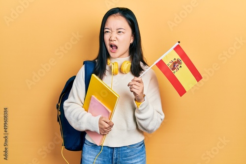 Young chinese girl exchange student holding spanish flag angry and mad screaming frustrated and furious  shouting with anger. rage and aggressive concept.