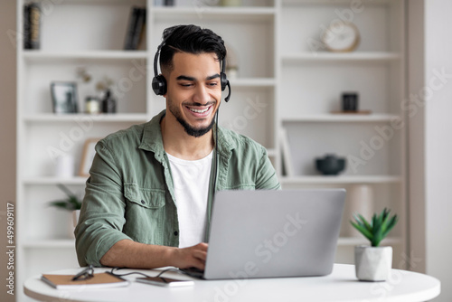 Happy handsome young islamic man with beard in headphones looks at laptop in living room interior