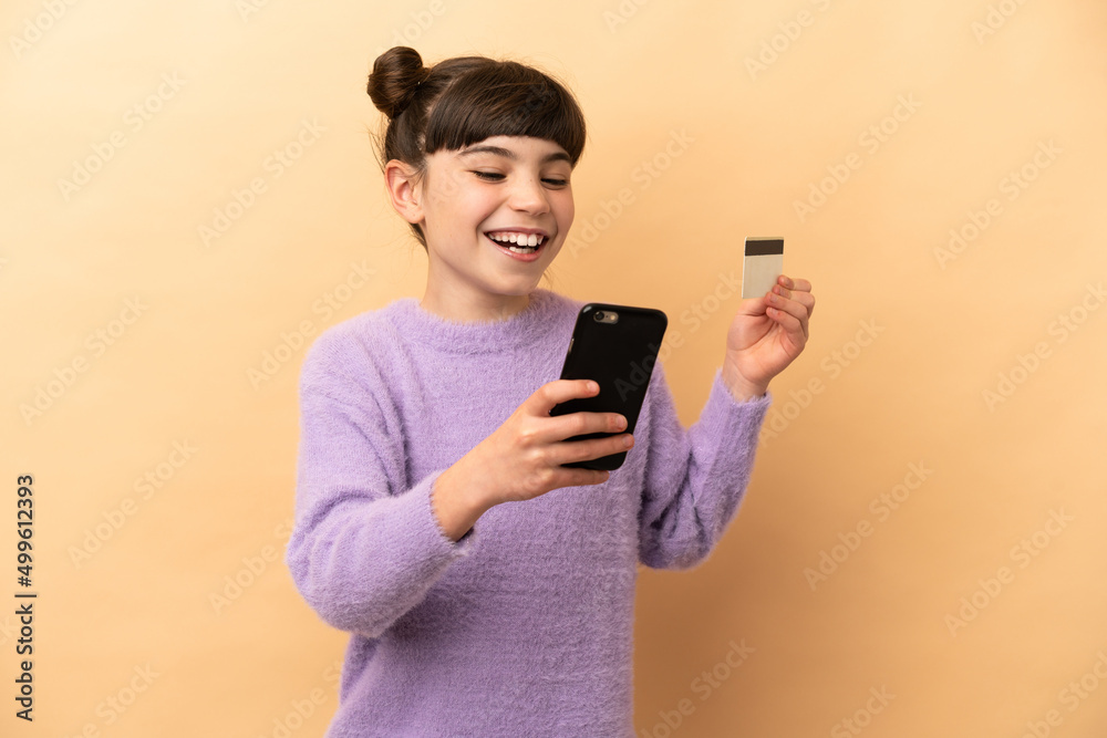 Little caucasian girl isolated on beige background buying with the mobile with a credit card Little caucasian girl isolated on beige background buying with the mobile with a credit card