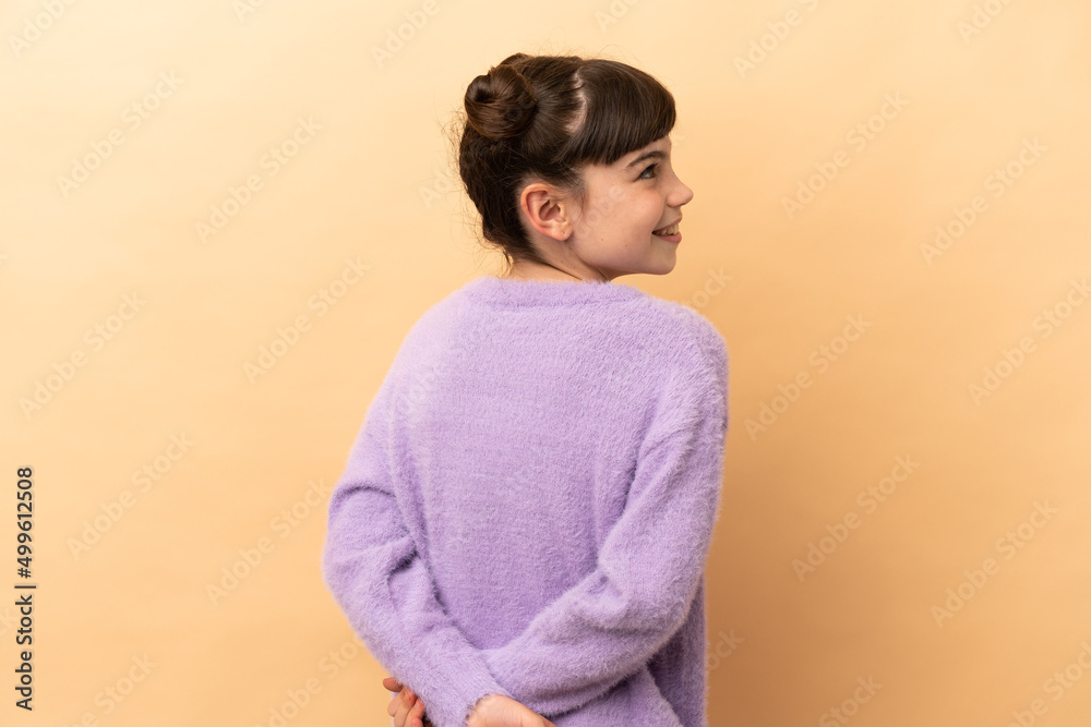 Little caucasian girl isolated on beige background in back position and looking back