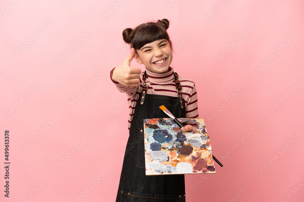 Little artist girl holding a palette isolated on pink background with thumbs up because something good has happened Little artist girl holding a palette isolated on pink background with thumbs up because something good has happened