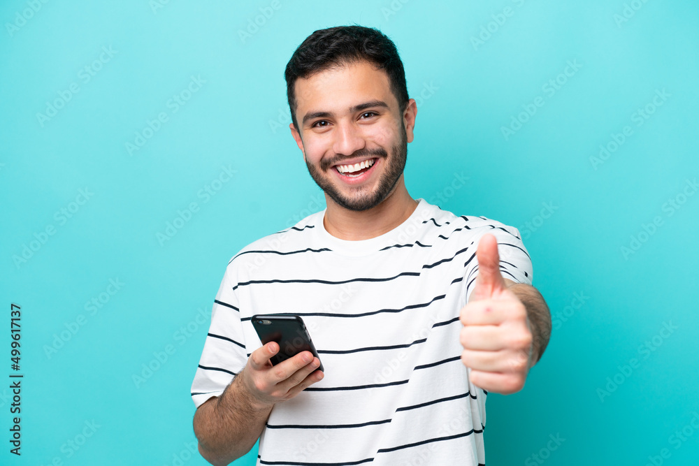 © luismolinero - Young Brazilian man isolated on blue background using mobile phone while doing thumbs up © luismolinero - Young Brazilian man isolated on blue background using mobile phone while doing thumbs up