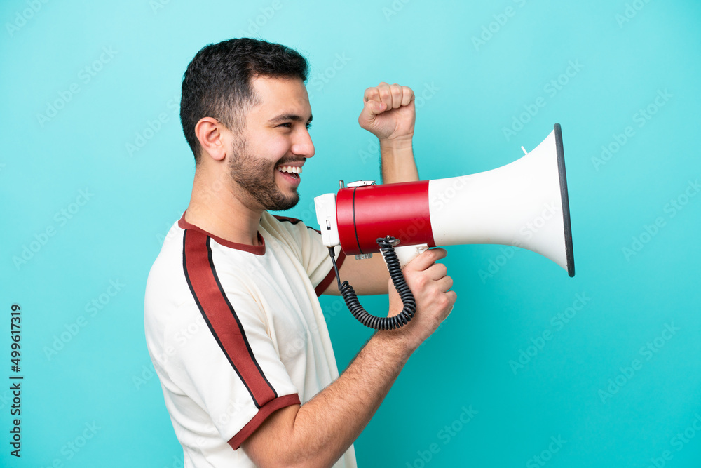 Young Brazilian man isolated on blue background shouting through a megaphone to announce something in lateral position Young Brazilian man isolated on blue background shouting through a megaphone to announce something in lateral position