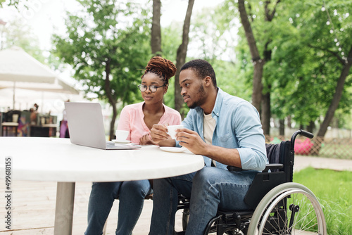 Young African American man in wheelchair and his female colleague using laptop together at table in outdoor cafe