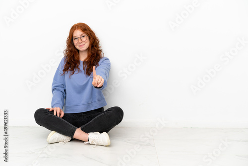 Teenager redhead girl sitting on the floor isolated on white background showing and lifting a finger