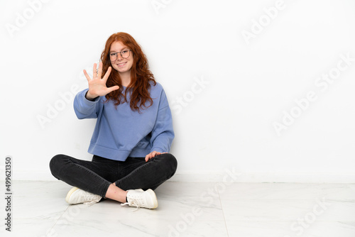 Teenager redhead girl sitting on the floor isolated on white background counting five with fingers
