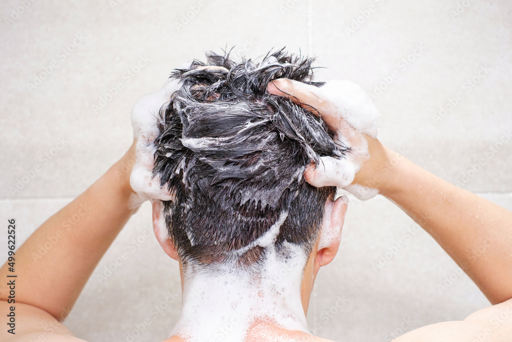 Male hand washes his head with shampoo and foam on gray background close-up, back view. - obrazy, fototapety, plakaty 