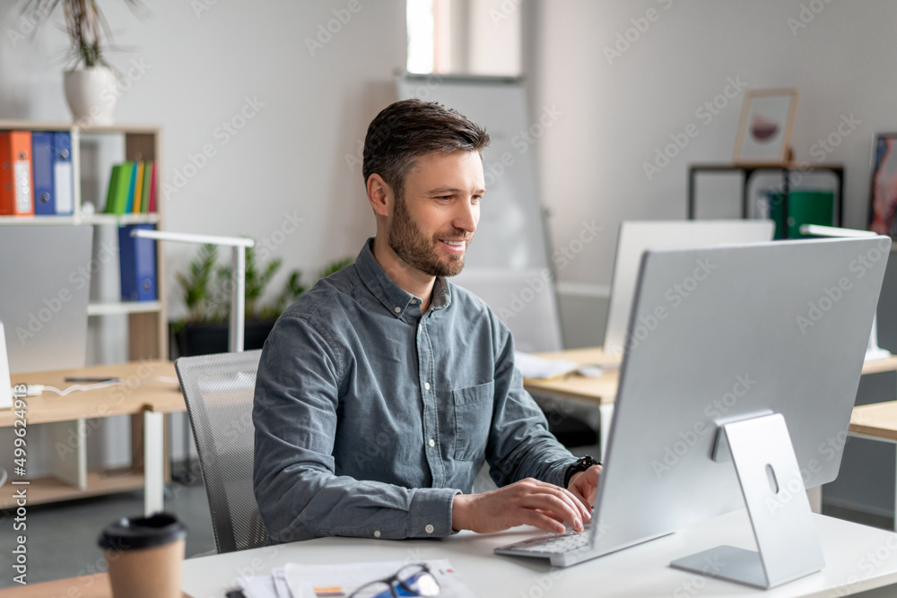 Cheerful middle aged businessman sitting at worktable, typing on computer keyboard, working on marketing research Cheerful middle aged businessman sitting at worktable, typing on computer keyboard, working on marketing research