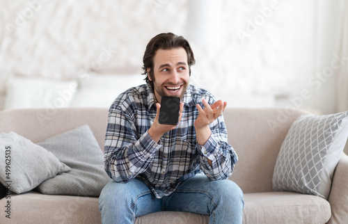 Cheerful young european man with stubble in casual talk by smartphone sits on sofa and gestures in living room
