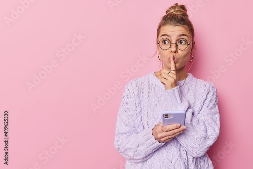 Mysterious young pretty woman shows shush gesture demands silence holds mobile phone sends text messages wears round spectacles and knitted sweater isolated over pink background mock up space