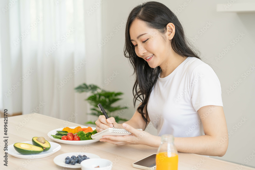 Diet, Dieting asian young woman write diet plan right nutrition on table with fresh vegetables salad, almond is different food ingredients in green. Nutritionist of healthy, nutrition of weight loss.