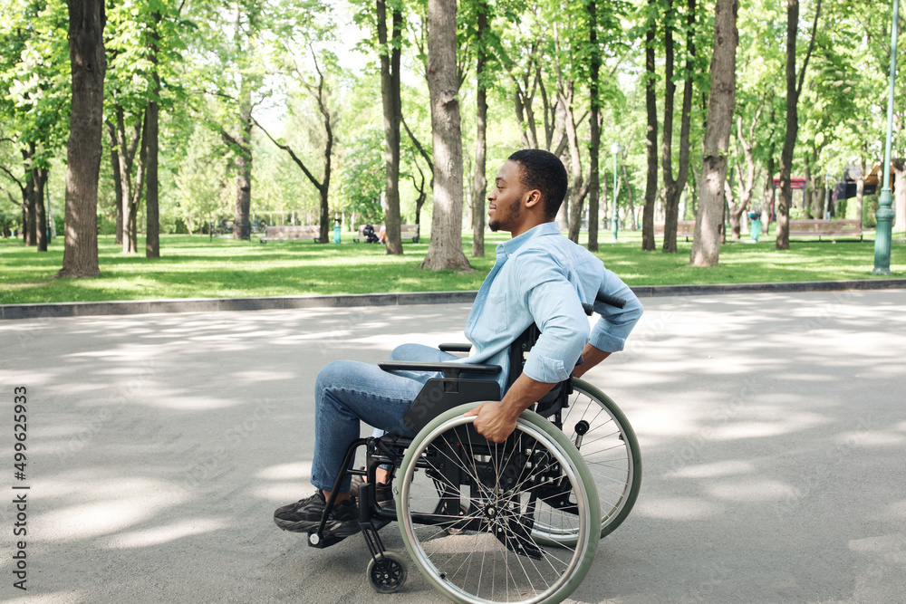 Side view of young black guy in wheelchair spending time at city park, full length Side view of young black guy in wheelchair spending time at city park, full length