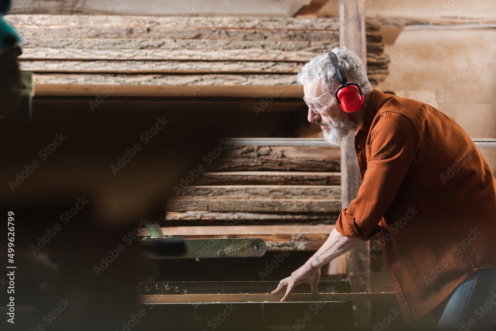 side view of carpenter in protective earmuffs working on circular saw on blurred foreground.