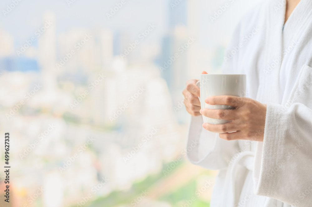 Side view of woman in bathrobe standing near the window while holding white cup of coffee and enjoy wonderful morning in the hotel, detaching from work. Side view of woman in bathrobe standing near the window while holding white cup of coffee and enjoy wonderful morning in the hotel, detaching from work.