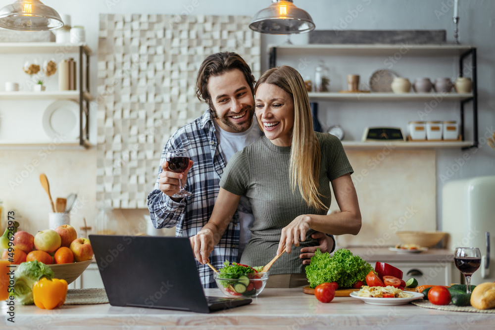 Satisfied millennial caucasian husband with stubble and wife cooking together, drink wine and talk by laptop Satisfied millennial caucasian husband with stubble and wife cooking together, drink wine and talk by laptop