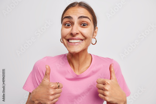 Glad brunette young woman smiles happily keeps thumbs up shows approval sign satisfied with service good feedback says its excellent dressed in casual pink t shirt isolated over white background