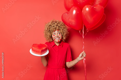 Positive woman has curly blonde hair wears festive dress holds bunch of heart balloons and sweet cake prepares for holiday isolated over vivid red background. People and celebration concept.