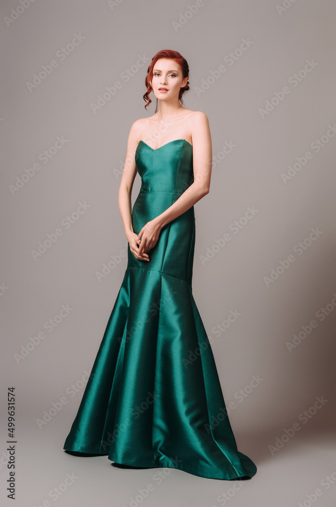 Emerald ball gown. Young lady in long green evening dress. Ginger woman posing in studio. Beautiful model wearing high heels, feminine look for an event. Women's classic fashion. Emerald ball gown. Young lady in long green evening dress. Ginger woman posing in studio. Beautiful model wearing high heels, feminine look for an event. Women's classic fashion.