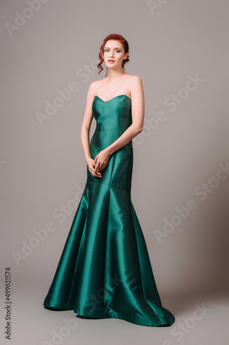 Emerald ball gown. Young lady in long green evening dress. Ginger woman posing in studio. Beautiful model wearing high heels, feminine look for an event. Women's classic fashion.
