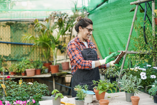 Mature latin woman working inside greenhouse garden using digital tablet