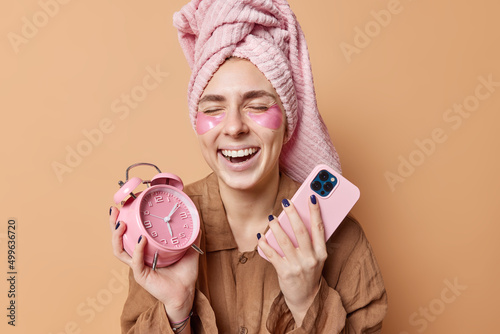 Cheerful woman has fun undergoes beauty procedures in morning applies hydrogel patches under eyes for moisturising delicate skin holds alarm clock showing time and modern smartphone dressed in pajama photo