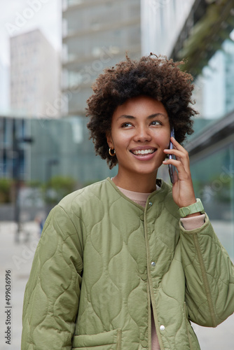 Vertical shot of happy curly haired woman talks via cellphone while walking in city smiles gladfully wears casual clothes being in good mood discusses latest news focused somewhere. Lifestyle concept