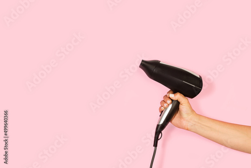 Mano de mujer sosteniendo un secador negro de pelo sobre un fondo rosa liso y aislado. Vista de frente y de cera. Copy space
