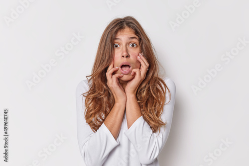 Omg concept. Speechless scared beautiful European woman with long hair keeps mouth opened stares bugged eyes reacts on shocking news keeps hands on face dressed casually isolated over white background