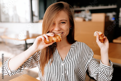 Excited charming lady with long light brown hairstyle is bitting a croissant and smiling while having breakfast in cafeteria