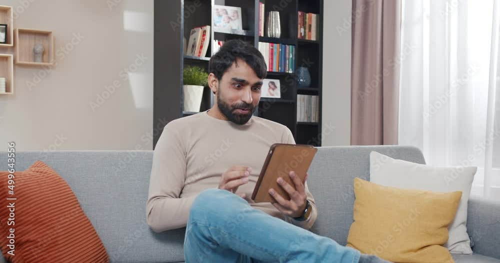 Arabian young handsome man sitting on couch in living room and using tablet device, is shocked and surprised happily. Happy young male getting good news online on computer at home. Arabian young handsome man sitting on couch in living room and using tablet device, is shocked and surprised happily. Happy young male getting good news online on computer at home.