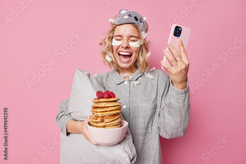 Joyful European woman has fun eats delicious pancakes with raspberries for breakfast holds mobile phone for sending text messages to friends in morning wears nightwear applies beauty patches