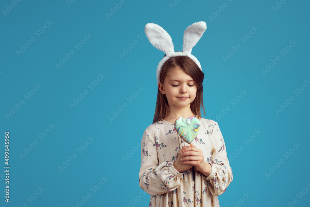 Horizontal photo of cute kid holding a cookie in shape of heart, wearing bunny ears and casual dress, isolated over blue background