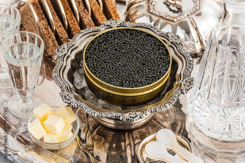 Black caviar on ice in silver bowl, Vodka and bread on white marble table close-up