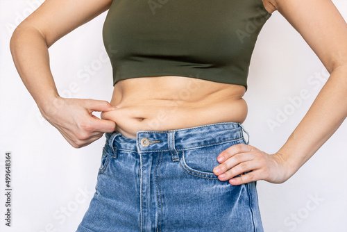 A cropped shot of a young woman in a short top and jeans holding herself by the fat on her stomach isolated on a white background. Overeating, excess weight concept