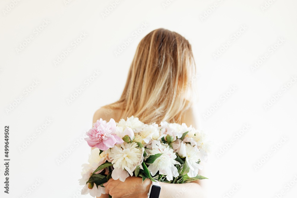 Conceptual portrait of young naked woman with blonde hair with bouquet of white peonies on beige background with copy space.