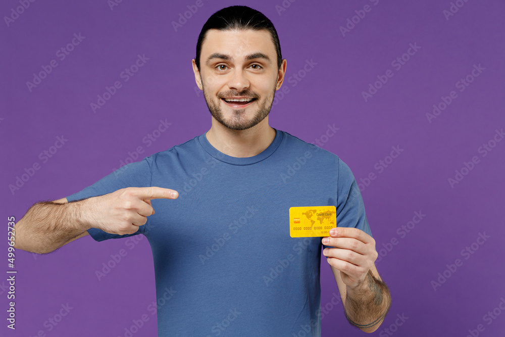 Young surprised amazed happy cheerful fun man 20s in basic blue t-shirt with blank print design hold point index finger on credit bank card isolated on plain purple color background studio portrait Young surprised amazed happy cheerful fun man 20s in basic blue t-shirt with blank print design hold point index finger on credit bank card isolated on plain purple color background studio portrait