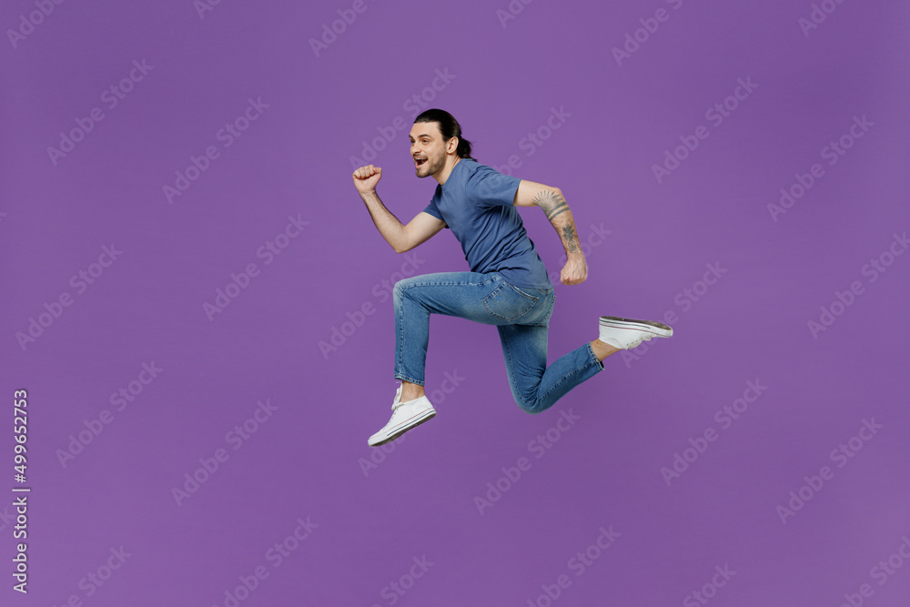 Full body side profile view young sporty man 20s wearing basic blue t-shirt jump high run fast in air hurrying up isolated on plain purple color background studio portrait. People lifestyle concept.