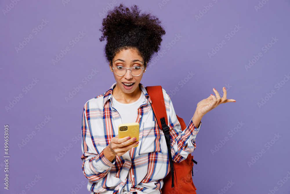 Young sad girl woman of African American ethnicity teen student in shirt backpack hold in hand use mobile cell phone isolated on plain purple background. Education in high school university concept. Young sad girl woman of African American ethnicity teen student in shirt backpack hold in hand use mobile cell phone isolated on plain purple background. Education in high school university concept.