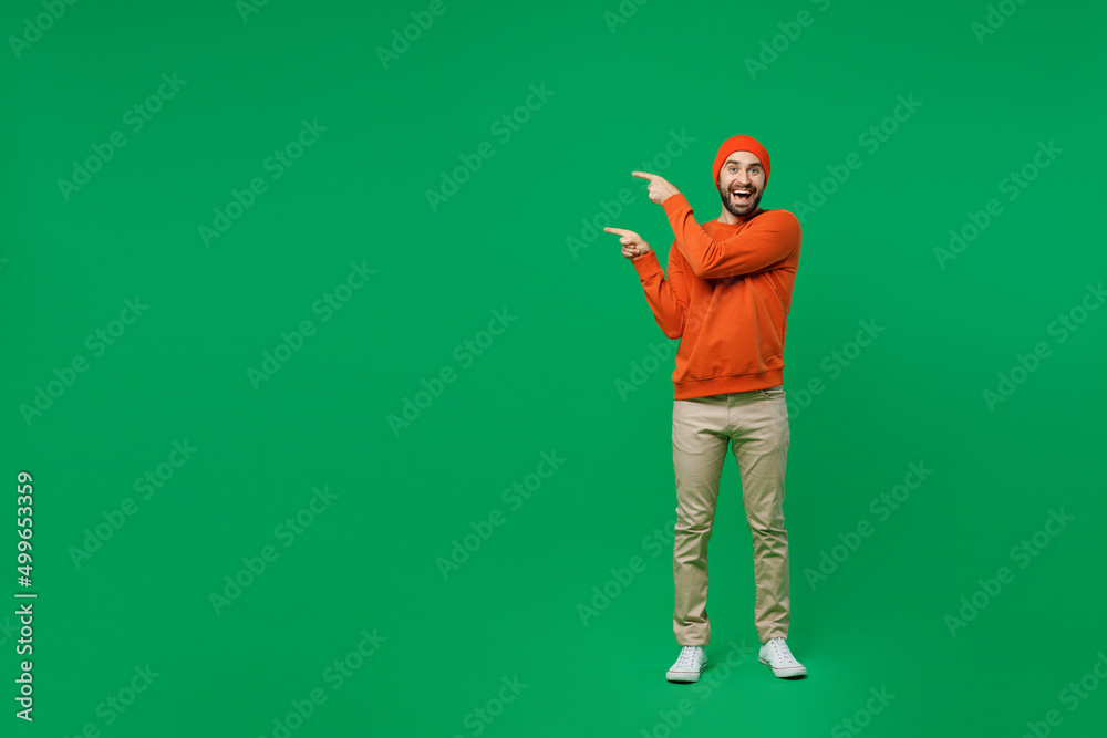 Full body young surprised happy amazed man 20s in orange sweatshirt hat point index finger aside on workspace area mock up isolated on plain green background studio portrait. People lifestyle concept. Full body young surprised happy amazed man 20s in orange sweatshirt hat point index finger aside on workspace area mock up isolated on plain green background studio portrait. People lifestyle concept.
