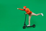 Full body side view young smiling happy fun man 20s wear orange sweatshirt hat riding electric scooter do super hero fly gesture isolated on plain green background studio. People lifestyle concept