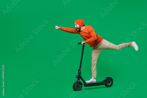 Full body side view young smiling happy fun man 20s wear orange sweatshirt hat riding electric scooter do super hero fly gesture isolated on plain green background studio. People lifestyle concept