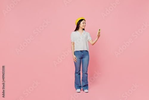 Full body smiling fun young woman of Asian ethnicity 20s wear white polka dot t-shirt yellow beret hold in hand use mobile cell phone isolated on plain pastel pink background People lifestyle concept