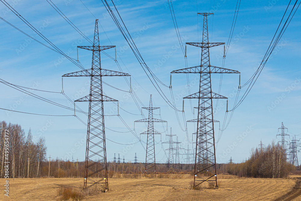 The towers of electric main in the countryside field on the background of blue sky with clouds and the forest with the wires in sunny or spring weather The towers of electric main in the countryside field on the background of blue sky with clouds and the forest with the wires in sunny or spring weather